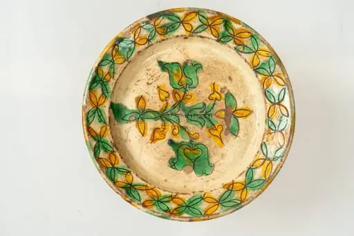 Farfurie de formă aplatizată, cu buză răsfrântă și muchie rotunjită. Piesa este modelată la roata olarului, angobată cu humă albă. Decorul este realizat prin sgrafitare în angoba crudă, peste care se aștern cu pensula culorile de smalț: verde și galben. Motivele decorative sunt florale și vegetale stilizate, dispuse pe fundul și pe buza farfuriei. Cromatica decorului: alb, galben, verde.