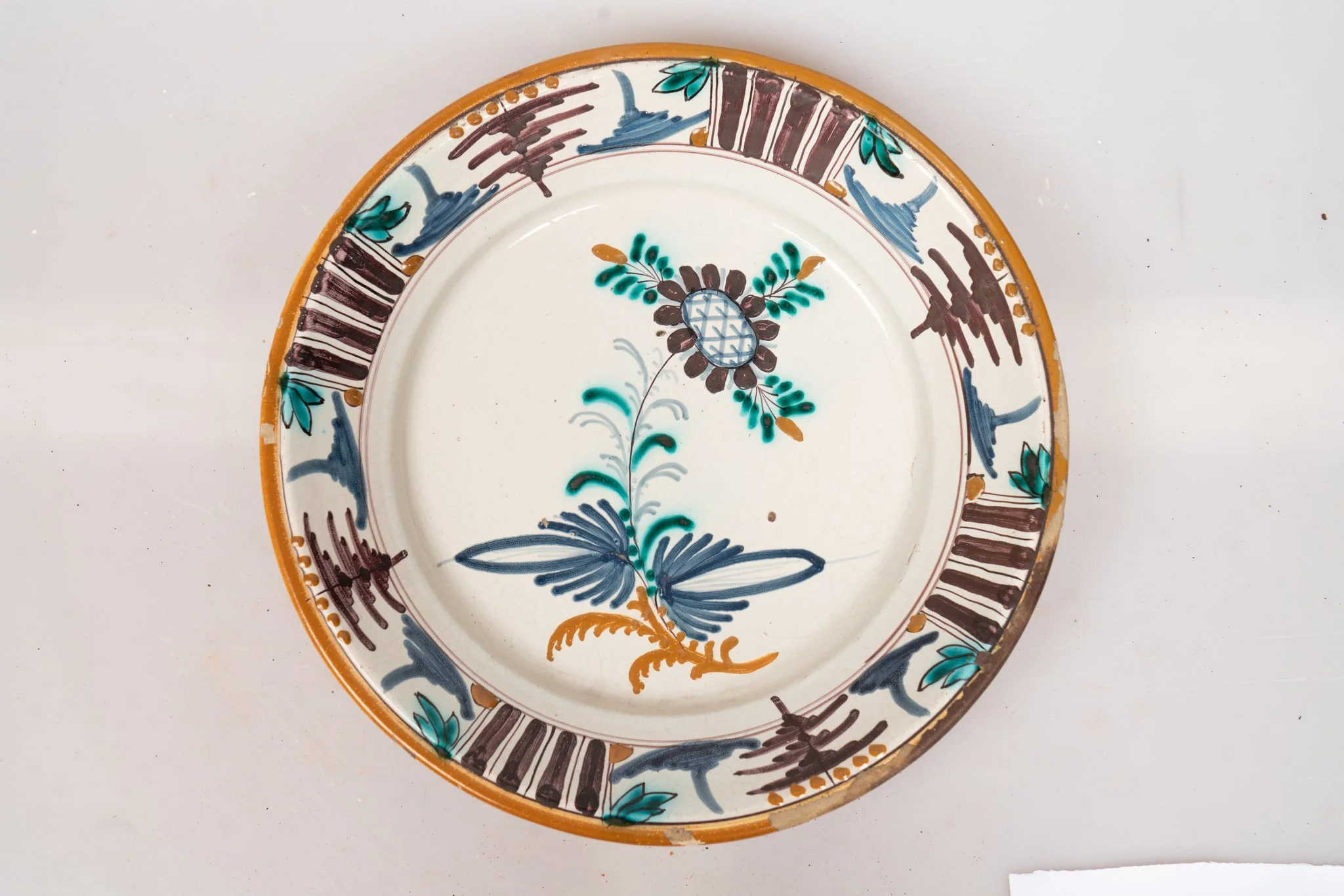 Farfurie de formă aplatizată, cu buza răsfrântă. Decorul, dispus pe un fond alb lăptos, prin pictare, este format dintr-un motiv floral stilizat, dispus central, pe fundul farfuriei, și motive geometrice (puncte galbene, grupuri de dungi paralele de culoare maro-brun, linii subțiri, paralele, linii sub formă de cerc, motivul bradului), dispuse într-un registru continuu pe buza farfuriei. Muchia buzei este marcată cu o dungă de culoare galbenă. Cromatica: alb, verde, brun, albastru, galben-ocru., by Unknown, 1850