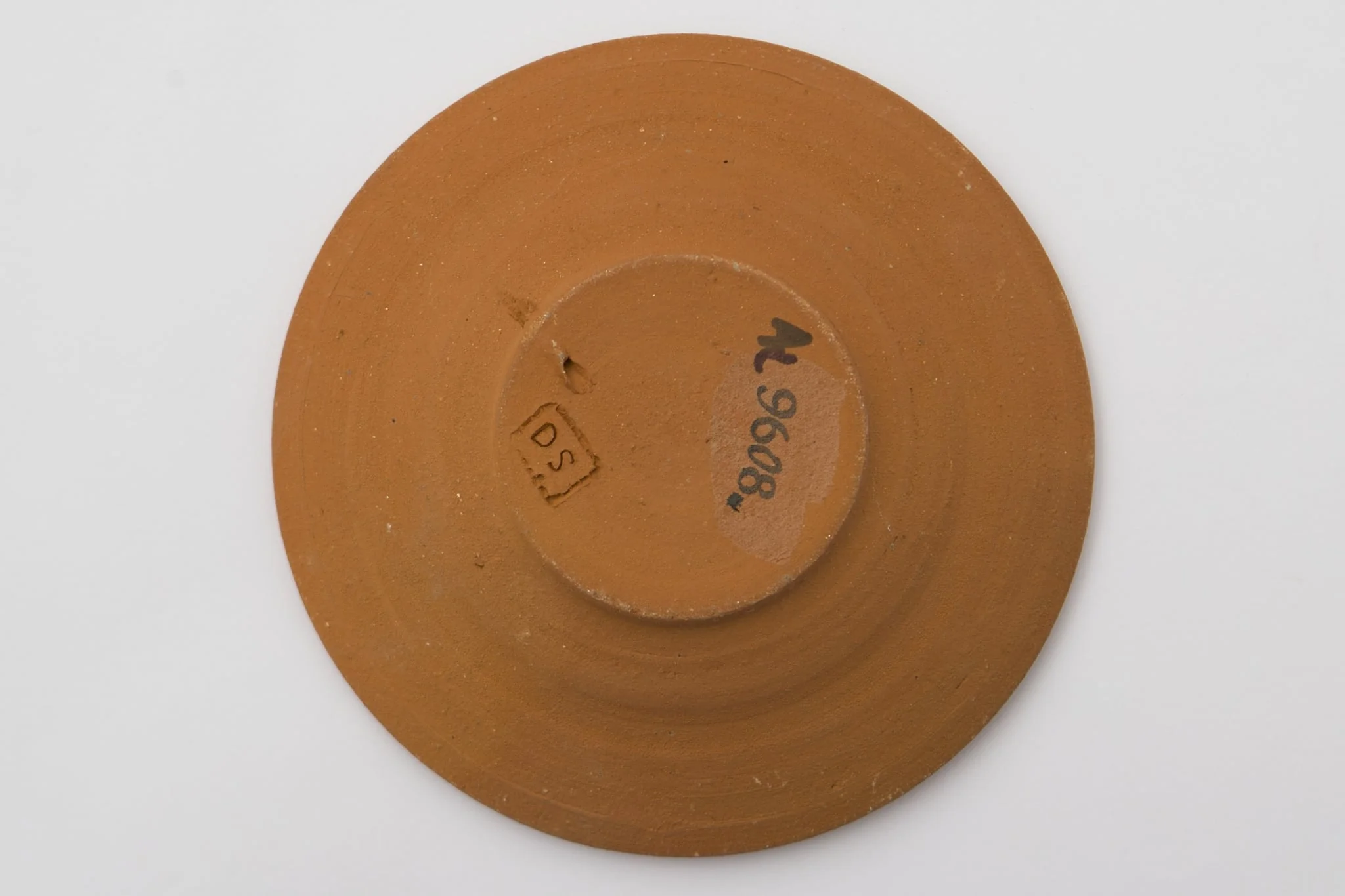 Vas ceramic cu fundul plat puțin adâncit și marginile evazate, puțin ridicate. Ornamentată cu pensula cu angoba albă. Fundul este marcat printr-o spirală din care ies alte șase spirale mici dispuse pe circumferința exterioară. Pe marginea vasului două benzi, una cu o linie întreruptă, a doua cu șase valuri întrerupte. La posterior ștanțat ”DS” încadrat într-un dreptunghi. Cromatică: fond: cărămiziu; motive: alb.
