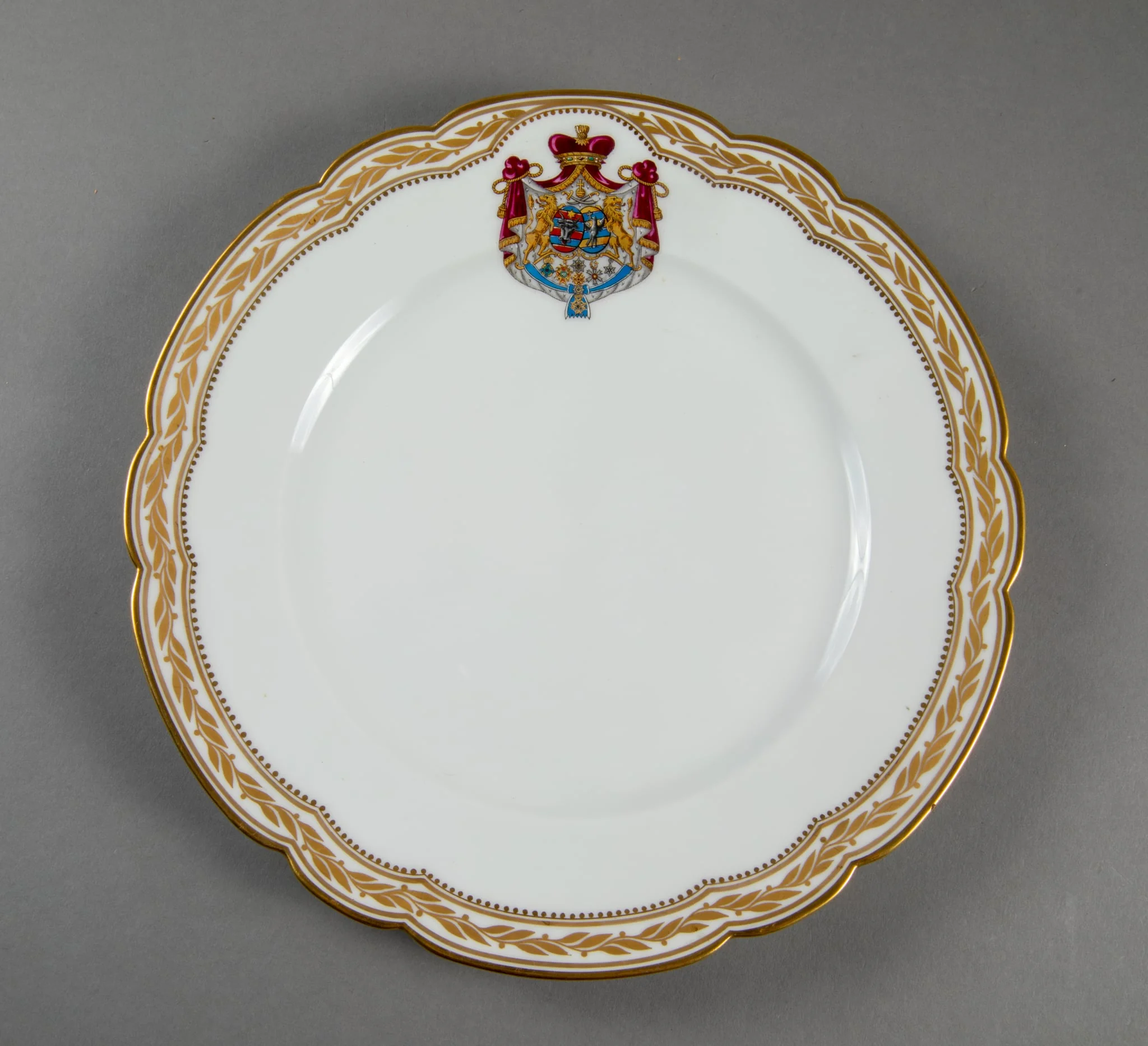 Farfurie de porțelan de Sevres, realizată din porțelan alb, cu margini treflate, decorate cu frunze de laur pictate, în centrul decorațiilor având blazonul familiei Suțu. Blazonul a fost adesea confundat cu cel al Principatelor Unite, fiind format din: stemele celor două țări românești: prima cea a Moldovei, cea de-a doua cea a Țării Românești, steme plasate pe două scuturi ovale, în fascie, acolate, puțin suprapuse, cea a Moldovei având capul de bour de argint cu stea în șase colțuri de aur, pe un scut cu segmente verticale egale roșii și albastre; cea a Țării Românești având acvila cruciată de argint, cu crucea de aur, scutul având segmente verticale galbene și albastre. Cele două scuturi au ca tenanți doi lei aurii, deasupra armele familiei Suțu – iataganul și sabia, ambele aurii, precum, pe un pavilion de purpură căptușit cu hermină, brodat cu ciucuri de aur și prins într-o coroană cu pietre scumpe. Pe scut, în partea inferioară se află și decorațiile familiei Suțu. Familia Suțu a fost o familie fanariotă grecească, venită în principate în cea de-a doua parte a secolului al XVIII-lea, având trei domni în ambele țări Românești (Alexandru Suțu, Mihai Suțu și Grigore Suțu). Blazonul familiei poate fi admirat inclusiv pe clădirea Palatului Suțu, astăzi Muzeul de Istorie a Municipiului București. Farfuria are marca atelierului Sevres și modelul din anul 1844, precum și seria., by Unknown, 1850