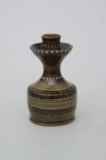 Recipient ceramic tubular, folosit ca suport de lumânare. Baza, fără fund, foarte înaltă cu pereții drepți ce se continuă printr-un gât îngust, înalt, lățindu-se în partea superioară pe marginea care se evazează la exterior. Este prevăzut un șanț pentru ținerea cerii topite. Partea superioară prevăzută cu un orificiu înalt pentru fixat lumânarea. Decorații jirăvite pe întreg corpul, linie verde ondulată pe umăr, linie punctiformă circulară la baza gâtului și pe inelul reliefat, și o linie în zig-zag în partea superioară a gâtului. Smălțuit în exterior. Cromatică: fond: maro închis; motive: alb; verde; maro; negru; bej.