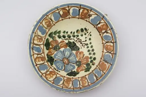 Recipient ceramic smălțuit, de formă tronconică, cu baza plată, pereții ușor ridicați și buza profilată. Peste stratul alb de angobă au fost realizate două registre ornamentate cu motive geometrizante și fitomorfe stilizate. Primul registru, situat pe fundul vasului este format din trei flori: una, centrală cu mijlocul maro și petalele albastre, celelalte două de culoare maro roșcat. Crenguțelor, pe care sunt poziționate florile, li se unesc vârfurile, deasupra cărora a fost trecută datare: „1960”. Marginal a fost realizat cel de-al doilea registru, format din boboci de culoare albastru și maro roșcat, despărțiți de linii verticale drepte. Două linii circulare drepte încadrează ultimul registru. Farfuria era utilizată în gospodărie pentru decorarea interioarelor. Prezintă uzură funcțională. Cromatică: fond: alb; motive: albastru; maro roșcat; verde.