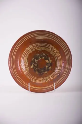 Farfurie de formă tronconică, marginea lată ușor înclinată în sus. Decor: pe fond de roșeală pe fundul plat, un cerc de puncte albe și negre în formă de coroniță; pe perete cercuri concentrice cu motive de linii verticale, pe margine trei cercuri concentrice albe. Smalț incolor. Cromatică: fond: maro; motive: alb-gălbui; negru.