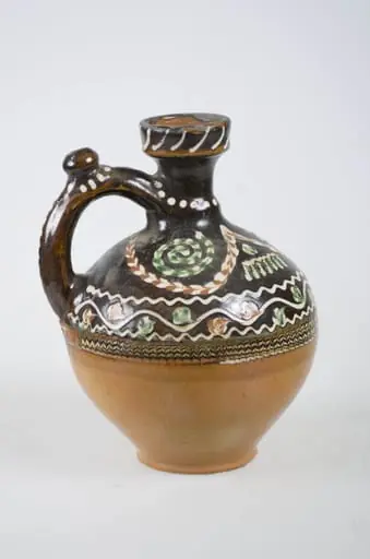 Vas ceramic sferic, ce prezintă baza plată din care pornesc pereții puternic arcuiți spre exterior, înalți, care în partea superioară se îngustează și se termină printr-un gât înalt cu marginea dreaptă ușor rotunjită. Ornamentul realizat cu cornul este dispus numai în partea superioară pe fond de angobă, realizat cu cornul. La diametrul maxim o porțiune jirăvită cu șarpe de o parte și de alta cu "altiță" și "boboci". Pe gât trei linii ondulate urmat de un șir de puncte. Pe marginea buzei linii. Mânerul prevăzut cu "țâță" are puncte. Jumătatea inferioară maro deschis. Smălțuit. Pe fundul vasului apare numele meșterului și localitatea "Ogrezeanu Stelian - Horezu". Cromatică: fond: maro închis partea superioară. Cărămiziu partea inferioară; motive: verde; alb; maro închis.