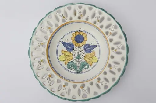 Farfurie ornamentală ce prezintă găurele între buză și partea adâncită obținute prin ștanțare. Buza este vălurită și prezintă un contur de culoare verde. Registrul central are motive florale și geometrice: cercuri galbene și albastre ce încadrează motivele florale. La posterior, baza inelară perforată pentru atârnare. Cromatică: fond: alb; motive: verde; galben; albastru; negru.