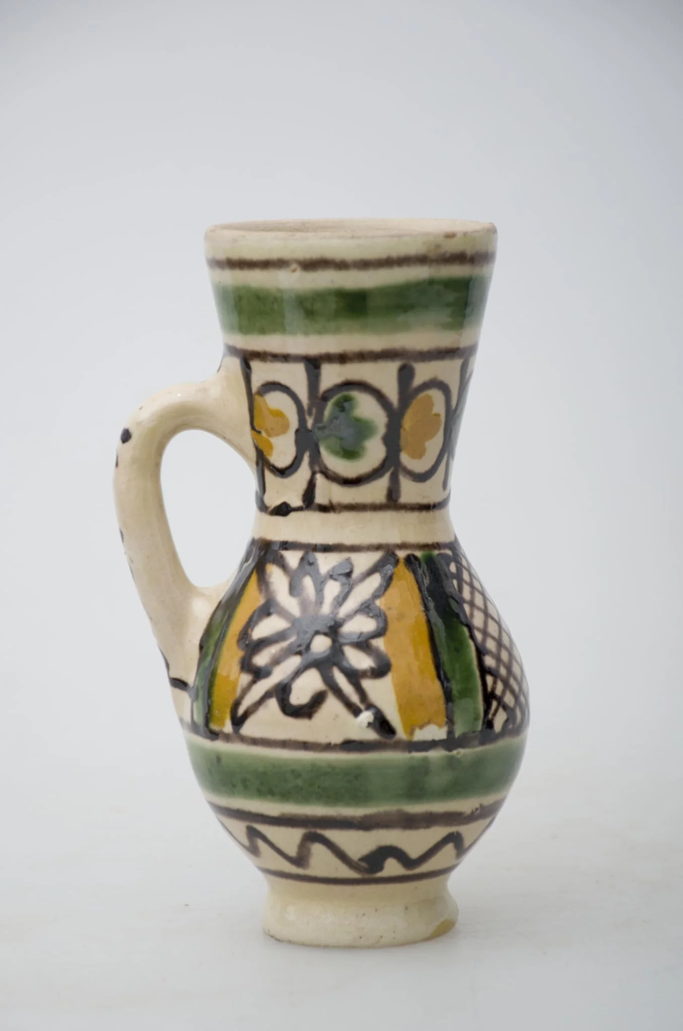 Vas ceramic de formă bitronconică, smălțuit de mici dimensiuni, cu baza plată și bombat în zona „pântecelui”. Prezintă o toartă ce pornește din zona gâtului redus ca dimensiuni și se termină în zona diametrul maxim al vasului. Decorul vasului este compus din două registre picturale. În primul registru, mărginit de două linii drepte subțiri, de culaore maro închis, au fost pictate două flori conturate cu negru și încadrate de câte două linii groase de culoare verde și galben. Cel de-al doilea registru este compus din boboci de culoare verde și galben, fiind dispuși concentric pe gâtul vasului. Ambele registre picturale sunt încadrate între două linii groase circulare, verzi. Pe toartă a fost redactat textul: „KORUND”. Prezintă uzură funcțională. Cromatică: fond: alb; motive: galben, verde, maro închis.