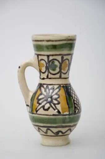 Vas ceramic de formă bitronconică, smălțuit de mici dimensiuni, cu baza plată și bombat în zona „pântecelui”. Prezintă o toartă ce pornește din zona gâtului redus ca dimensiuni și se termină în zona diametrul maxim al vasului. Decorul vasului este compus din două registre picturale. În primul registru, mărginit de două linii drepte subțiri, de culaore maro închis, au fost pictate două flori conturate cu negru și încadrate de câte două linii groase de culoare verde și galben. Cel de-al doilea registru este compus din boboci de culoare verde și galben, fiind dispuși concentric pe gâtul vasului. Ambele registre picturale sunt încadrate între două linii groase circulare, verzi. Pe toartă a fost redactat textul: „KORUND”. Prezintă uzură funcțională. Cromatică: fond: alb; motive: galben, verde, maro închis.
