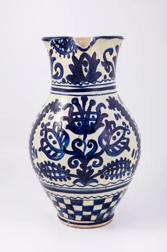 Cană piriformă prevăzută cu o toartă dreaptă și lată, gura rotundă, prevăzută cu cioc de scurgere. Suprafața decorativă a vasului este împărțită în patru registre decorative. Primul registru dispus pe gâtul piesei prezintă un decor floral și fitomorf reprezentând lalele și lujeri. Al doilea registru prezintă o linie vălurită și trei linii subțiri circulare. Decorul central, reprezintă trei „arbori ai vieții” stilizați realizați din trei lalele mari, înflorite, lujeri și frunze, separați de câte o semilună. Registrul al patrulea prezintă motivul tablei de șah. Deasupra toarte pictate cu motive florale și fitomorfe este redat anul confecționării piesei: 1980, iar pe fundul vasului numele persoanei care a pictat piesa: Herta Wilk. Cromatica: alb, albastru-cobalt.