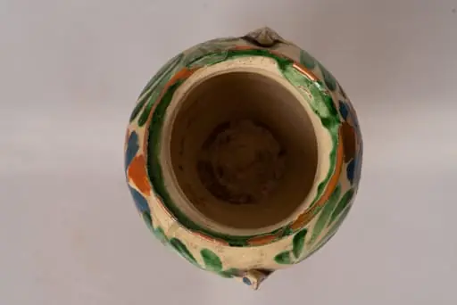 Piesă cu corp discoidal bombat, cu doi butoni triunghiulari aplicați pe corpul vasului în zona mediană și decorați cu puncte pictate. Buza vasului este marcată cu o linie pictată iar gâtul de un șir de puncte de culoare maro și albastru. Corpul vasului este decorat cu motive florale (floare rotundă cu opt petale) și motive fitomorfe, caracteristice centrului: frunze alungite de culoare verde. Sub decorul central apar linii concentrice pictate cu cornul, de culoare verde. Cromatica: alb, verde, maro, albastru. Vasul este smaălțuit transparent pe ambele fețe.