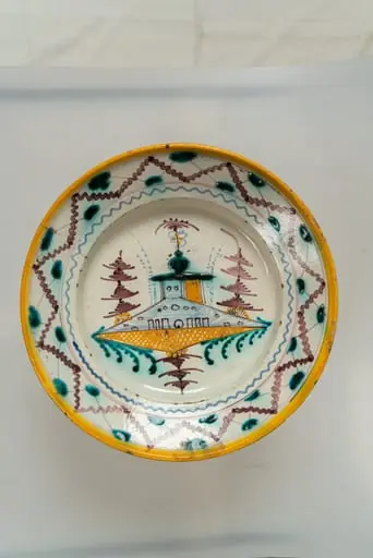 Farfurie de formă aplatizată, cu marginea lată, răsfrântă. Decorul principal este dispus în centrul farfuriei, pe un fond alb-lăptos, și prezintă un motiv arhitectural și vegetal stilizat, iar buza farfuriei este decorată cu motive geometrice (două linii vălurite dispuse în zig-zag, alternativ, una mai groasă, cealaltă mai subțire, de culoare maro; pete de culoare verde turcoaz, linii subțiri dispuse sub formă de cercuri concentrice). Muchia buzei este marchată de o dungă galbenă. Cromatica: alb, maro, galben ocru, albastru deschis, verde turcoaz.
