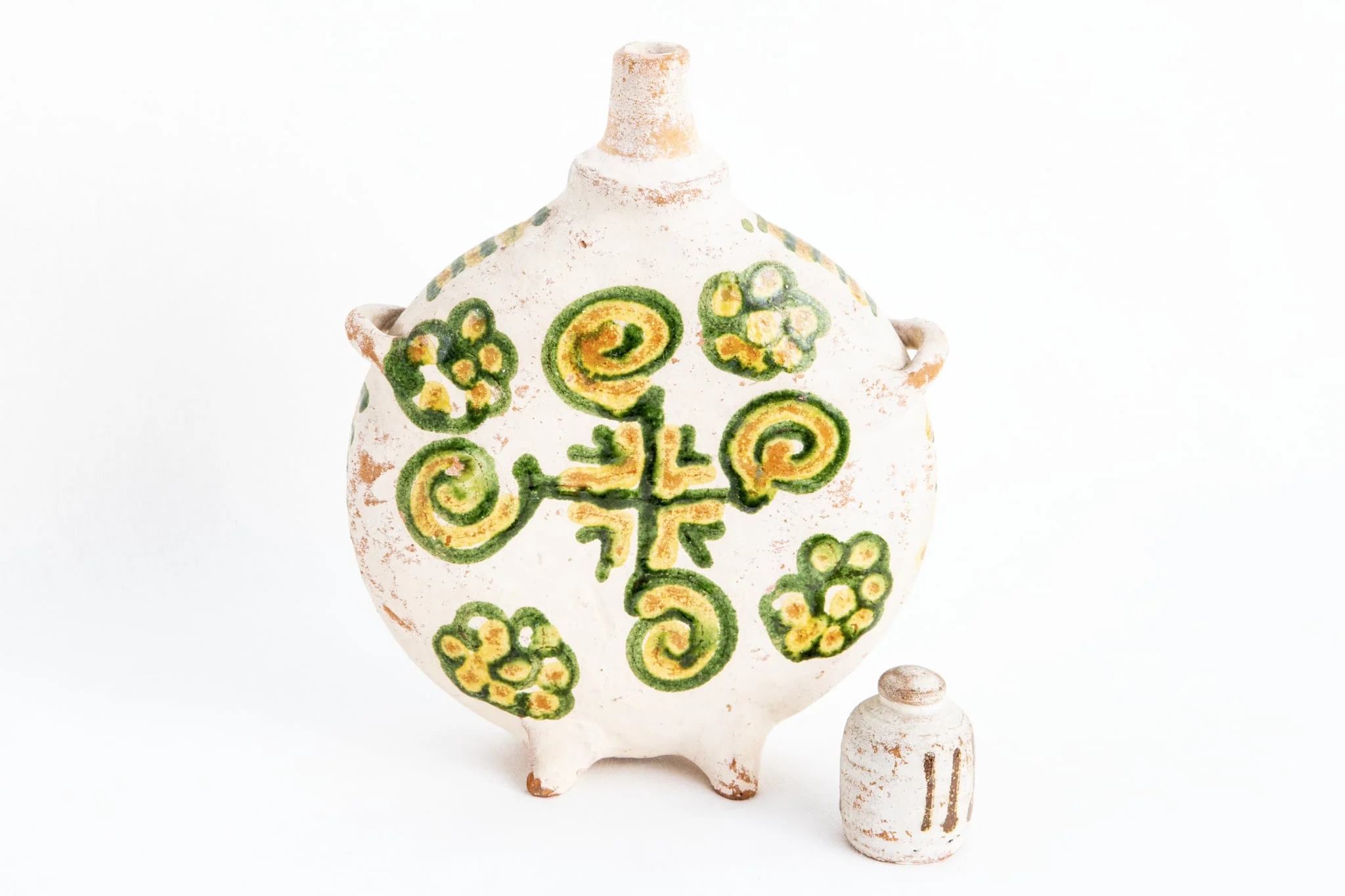 Ploscă de dimensiuni mici prevăzută cu capac. Fond alb. Decorul este similar pe ambele fețe ale obiectului; o cruce de culoare verde dispusă central; între brațele crucii sînt amplasate unghiuri duble colorate în galben respectiv, verde. Crucea este încadrată de patru spirale colorate în galben și verde și patru flori stilizate realizate cu aceleași culori. Muchia ploștii este decorată cu linii orizontale de culoare galbenă și verde, în alternanță cromatică., by Dumitru Șchiopu