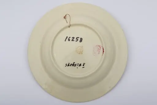 Piesă ceramică smălțuită, de formă circulară, cu baza plată, pereții puțin înălțați și marginile larg evazate. Peste stratul alb de angobă a fost pictat decorul fitomorf stilizat. Central, pe fundul piesei au fost realizați trei trandafiri roșii, însoțiți de patru boboci roșii și frunze verzi. Primul registru este încadrat de cel de-al doilea registru, dispus pe marginea recipientului ceramic. O bandă circulară galbeană peste care au fost realizați șase boboci roșii și frunze maro și verzi, este mărginită de două linii concentrice drepte. Farfuria era folosită în gospodărie pentru servirea hranei sau amenajarea interioarelor. Prezintă uzură funcțională. Cromatică: fond: alb; motive: roșu; verde; galben; maro.