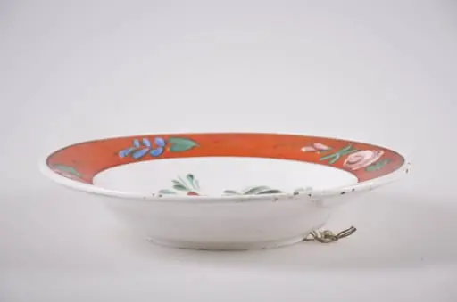 Vas de formă tronconică din faianță, cu buza dreaptă, ornamentată prin pictare. Fundul alb, pe care sunt pictate trei flori roșii (bujor) înconjurate de frunze mărunte, este delimitat de buza farfuriei cu o linie subțire, neagră. Buza vasului este roșie, decorată cu flori roz și frunzulițe discrete verzi și albastre. În partea posterioară prezintă o gaură prin care se trece ața ce servește la agățarea farfuriei. Cromatică: fond: alb; motive: roșu; verde; roz; albastru; negru.