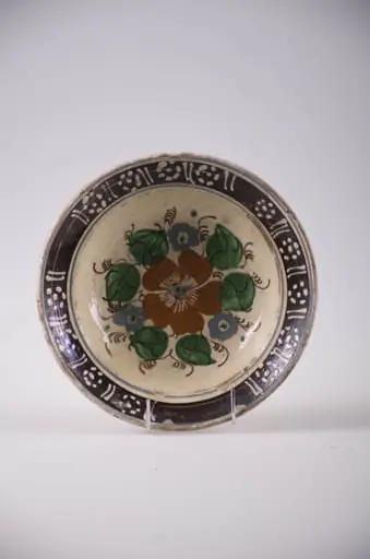 Blid de formă tronconică cu partea superioara usor rasfrântă, Decorul este pictat cu pensula cu un motiv central de tip floral; pe margini linii paralele și flori stilizate din puncte. Smălțuit. Cromatică: roșu; verde și maro în centru; motive laterale: alb pe fond brun inchis; fond general: alb-galbui.