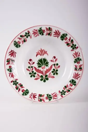 Farfurie de formă tronconică, adâncită; pe fund are găuri pentru agățare, Este decorată cu motive florale și geometrice; pe margine este decorat cu flori încadrate la exterior de o linie; în centru are mai multe flori. Pe fund are o marcă indescifrabilă. Cromatică: fond: alb; motive: vișiniu; verde.