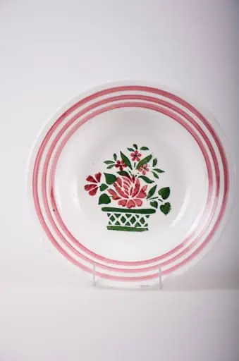 Vas de formă tronconică, adâncit; pe fund are găuri pentru agățare. În centru este decorată cu motive florale: vază cu flori; pe margine sunt trei cercuri concentrice; motive geometrice: linie, romb, triunghi. Pe fund are o marcă indescifrabilă. Cromatică: fond: alb; motive: vișiniu; verde.