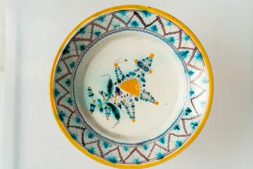 Farfurie mare, de formă aplatizată, cu marginea lată, răsfrântă, și cu muchia buzei ușor concavă spre exterior. Decorul, dispus pe un fond alb, prin pictare, este format din motive florale fin stilizate (flori și frunze), dispuse central, pe fundul farfuriei, și motive geometrice pe buza acesteia. Cromatica decorului: alb, verde-turcoaz, brun, galben-ocru, albastru.