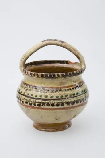 Vas ceramic de mici dimensiuni, pe bază inelară, pântecos, cu fundul și gura late. Deasupra gurii vasului a fost pusă o toartă de prindere. Buza vasului a fost pictată printr-un brâu de crenguțe. Pe pântecul vasului brâuri de puncte și două benzi subțiri jirăvite. Smălțuit în interior și exterior. Cromatică: fond: crem; motive: verde; maro; bej.