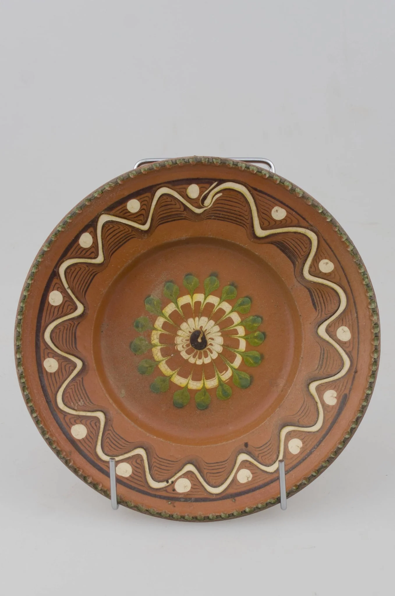Farfurie cu pereții rotunjiți, marginea oblică lată și buza crestată. Prezintă decor geometric pe margine: linii frânte și curbe; pete de culoare; central avem o floare stilizată. Cromatică: fond: maro; motive: verde; alb., by Răducanu Ion