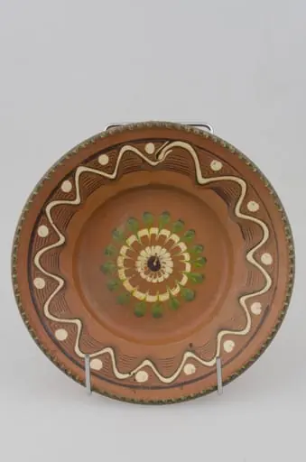 Farfurie cu pereții rotunjiți, marginea oblică lată și buza crestată. Prezintă decor geometric pe margine: linii frânte și curbe; pete de culoare; central avem o floare stilizată. Cromatică: fond: maro; motive: verde; alb.