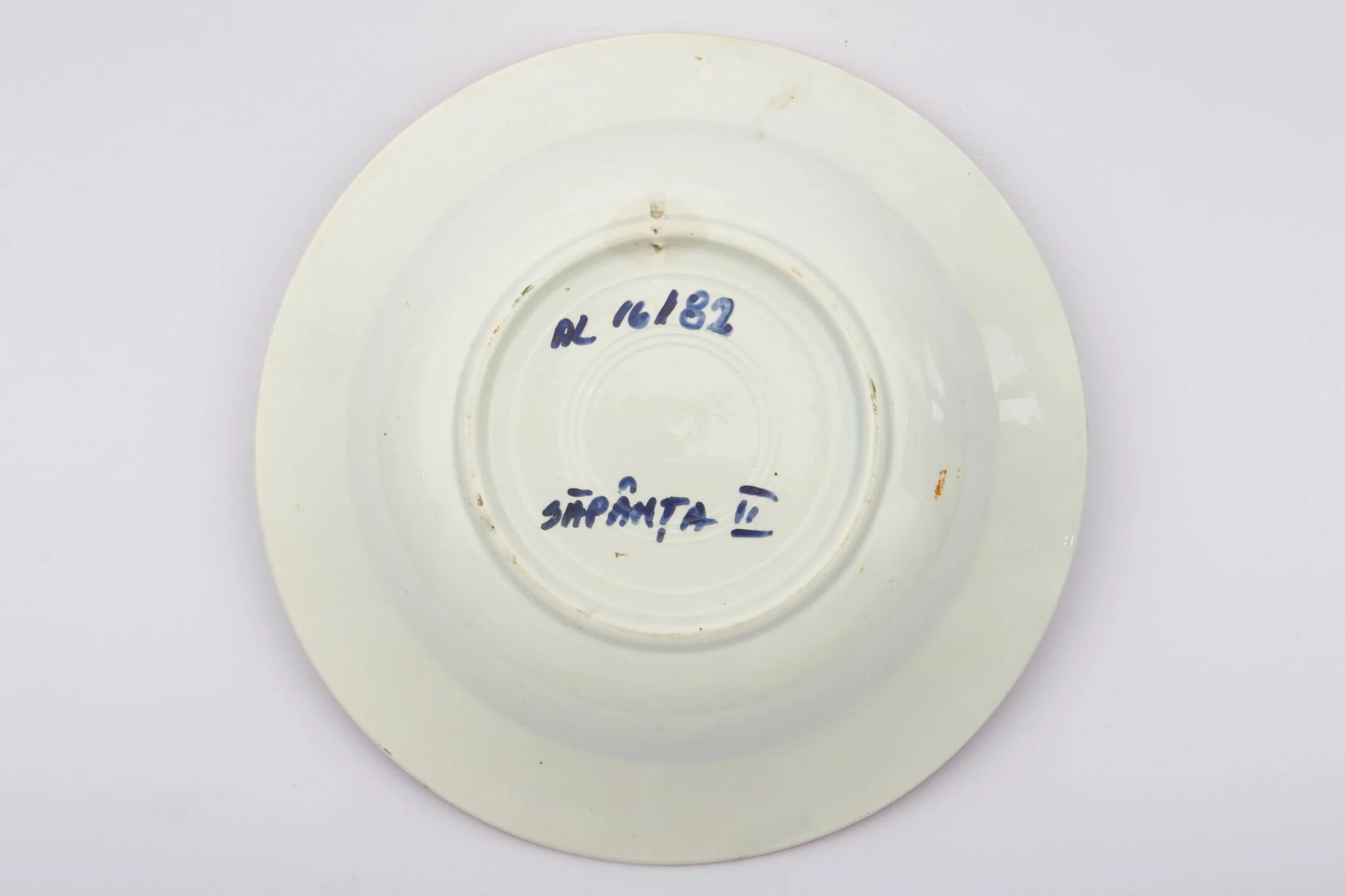 Vas ceramic de dimensiuni reduse, având formă tronconică, cu baza plată, pereții reficați și marginile evazate. Peste fondul alb au fost realizate decorațiile geometrizante și fitomorfe stilizate, dispuse în două registre. Primul registru este format dintr-un trandafir portocaliu cu frunze verzi, având în laterale câte o floare cu mijlocul albastru și petalele roșii, și câte un boboc roșu. Al doilea registru este format dintr-o tulpină circulară verde, pe care se dezvoltă trei grupuri de flori mici albastre. Alte trei grupuri, a câte trei linii portocalii oblice, însoțesc decorul fitomorf. Acest ultim registru este încadra dinspre interior de bandă groasă portocalie. Buza farfuriei a fost la rândul ei conturată cu portocaliu. Prezintă uzură funcțională. Cromatică: fond: alb; motive; portocaliu; verde; albastru., by Unknown, unspecified, 1850