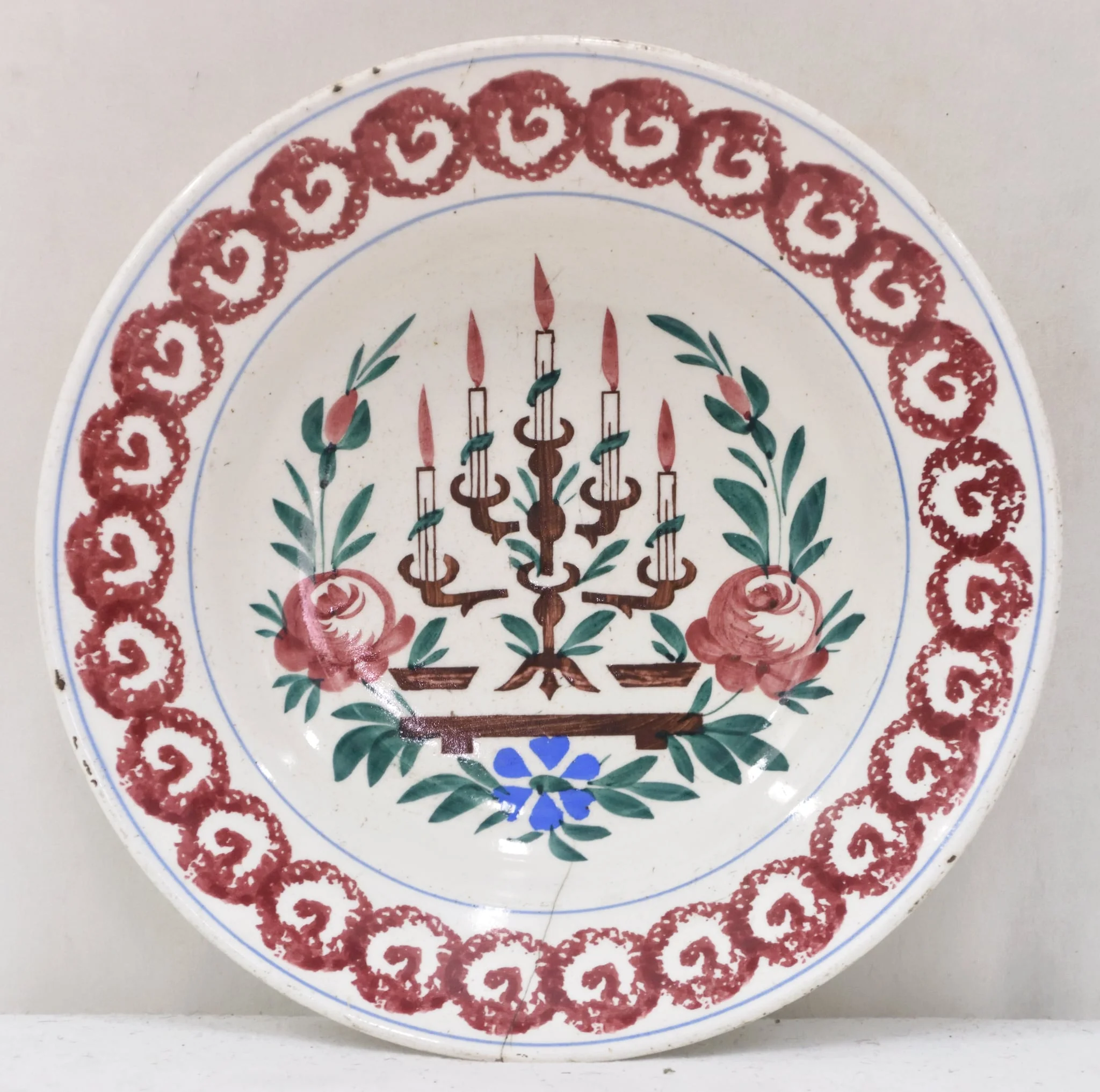 Obiectul are o formă tronconică cu baza mică în partea inferioară. Decorul se află numai la interiorul farfuriei fiind pictat cu motive geometrice, florale, vegetale și un simbol religios: pe fundul farfuriei a fost pictată o menoră cu cinci brațe (sfeșnic - obiect de cult evreiesc) sprijinită pe un suport; fiecare dintre brațele sfeșnicului susține câte o lumânare aprinsă; această reprezentare este încadrată de tulpini, frunze, flori și boboci (trei flori plus doi boboci); întreaga pictură de pe fundul farfuriei este încadrată de un cerc iar un altul este "trasat" de jur-împrejur în partea superioară, la buza piesei; între cele două cercuri se găsește un șirag format din cercuri (cu diametre egale) tangente la exterior, asemănător unui lanț cu zale rotunde; fiecare dintre aceste cercuri are la interior un semn, ce amintește de litera "V". Pe spatele farfuriei se află un mic orificiu rotund prin care se introduce firul textil pentru posibilitatea atârnării obiectului de perete. Culoarea piesei este albă iar pictura este realizată cu următoarele culori: maro închis, maro deschis, bleu și verde., by Unknown, 1850