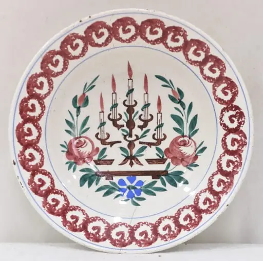 Obiectul are o formă tronconică cu baza mică în partea inferioară. Decorul se află numai la interiorul farfuriei fiind pictat cu motive geometrice, florale, vegetale și un simbol religios: pe fundul farfuriei a fost pictată o menoră cu cinci brațe (sfeșnic - obiect de cult evreiesc) sprijinită pe un suport; fiecare dintre brațele sfeșnicului susține câte o lumânare aprinsă; această reprezentare este încadrată de tulpini, frunze, flori și boboci (trei flori plus doi boboci); întreaga pictură de pe fundul farfuriei este încadrată de un cerc iar un altul este "trasat" de jur-împrejur în partea superioară, la buza piesei; între cele două cercuri se găsește un șirag format din cercuri (cu diametre egale) tangente la exterior, asemănător unui lanț cu zale rotunde; fiecare dintre aceste cercuri are la interior un semn, ce amintește de litera "V". Pe spatele farfuriei se află un mic orificiu rotund prin care se introduce firul textil pentru posibilitatea atârnării obiectului de perete. Culoarea piesei este albă iar pictura este realizată cu următoarele culori: maro închis, maro deschis, bleu și verde.