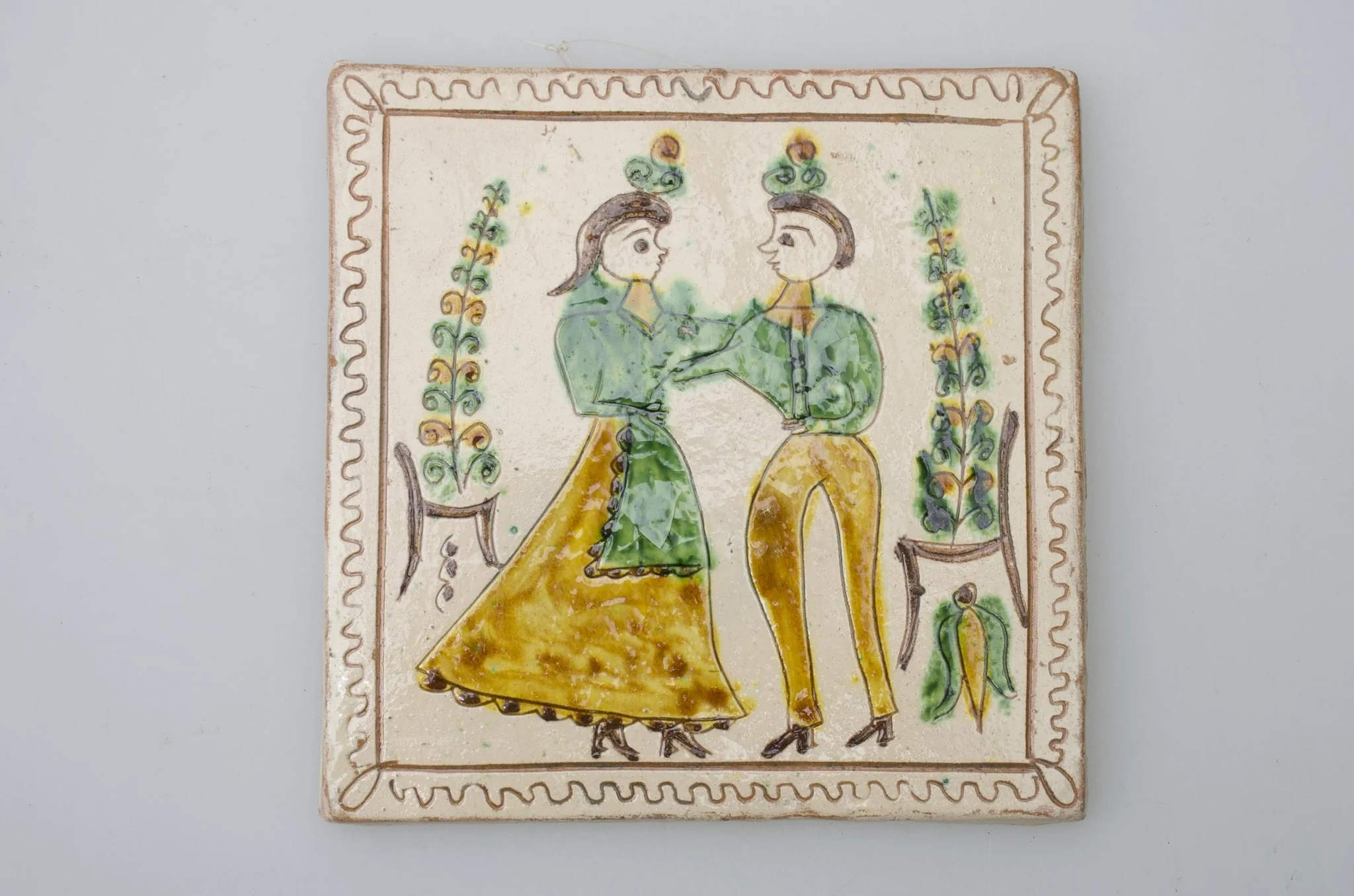 Piesă ceramică de formă pătrată, smălțuită și angobată, cu rol decorativ. Laturile pătratului sunt marcate de două chenare, primul vălurit, al doilea în linie dreaptă. Ornamentată cu motive antropomorfe și floral-vegetale: doi tineri, un băiat și o fată ținându-se pe după mijloc (băiatul cu mâna dreaptă, fata cu stânga). În spatele lor câte un scaun din care se înalță o floare. Motivele au fost la început incizate, iar apoi pictate. Pe spate, cahla este prevăzută cu un orificiu pentru prinderea aței. Tot aici, este inscripționat numele meșterului olar, "Colibaba". Cromatică: fundal alb; motive: verde, galben, cărămiziu, maro, negru.