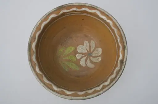 Farfurie adânca, de formă tronconică, smălțuită în interior. Pe margine prezintă decor geometric: linia vălurită; central o floare. Cromatică: fond: cărămiziu; motive: alb; verde; albastru.