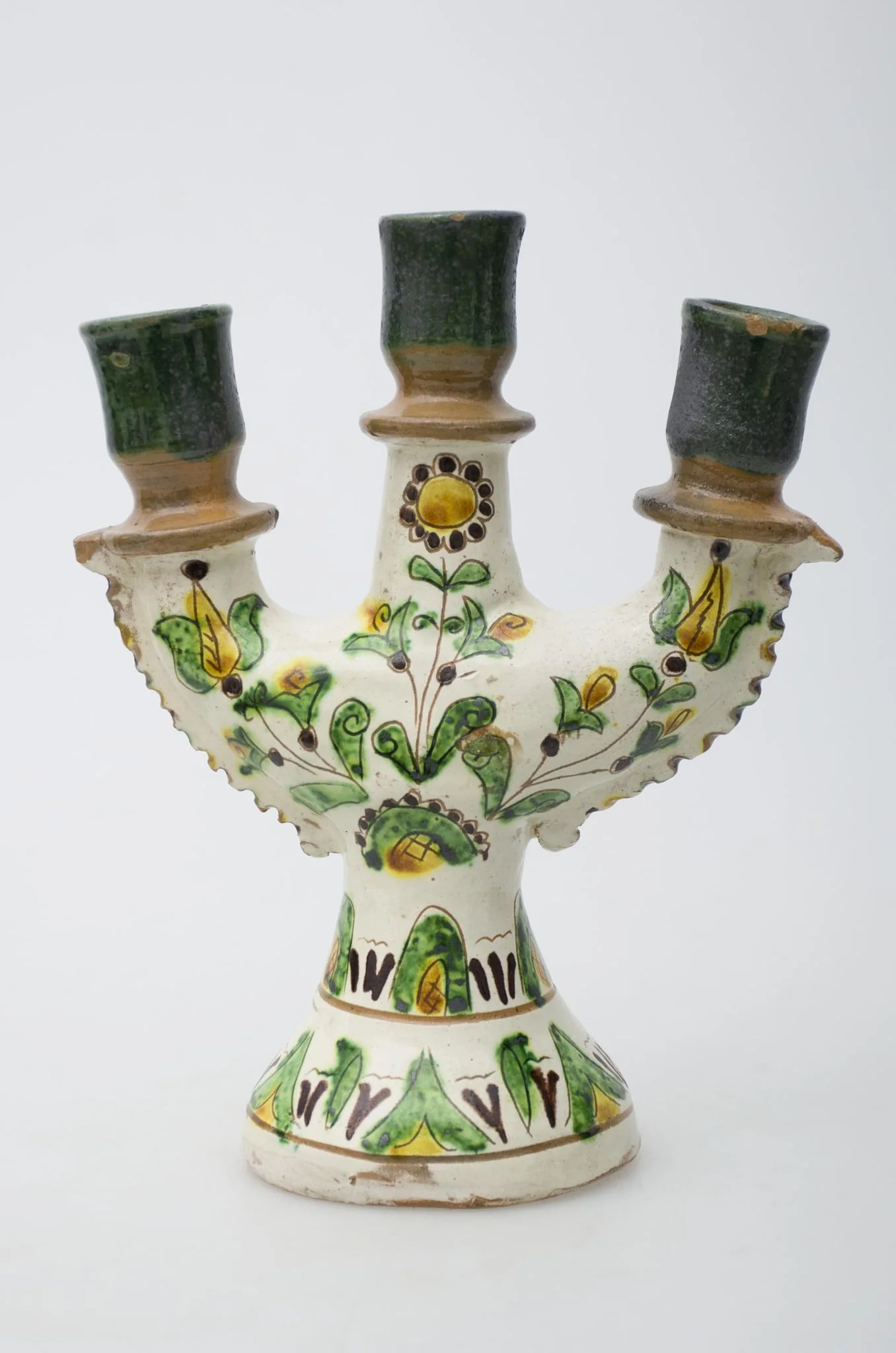 Obiect ceramic de uz casnic, format dintr-un picior tronconic, din care pornesc trei brațe, în care se introduc lumânările. Decorul pictural, realizat pe toată suprafața obiectului, este compus din motive geometrizante și vegetale. Primul registru, situat pe baza evazată, este format din elemente vegetale, dispuse circular și încadrat între două linii concentrice subțiri. Cel de-al doilea registru prezintă decor similar celui din registru inferior. Registrul superior este compus din trei flori, dintre care două (laterale) cu pistilul pronunțat, cea de-a treia, poziționată central, floarea soarelui. Gâtul și buza vasului au fost decorate cu verde și maro deschis. Iscălitură: indescifrabil. Cromatică: fond: alb; motive: crem; verde, maro închis, maro deschis, galben, negru., by Unknown, unspecified, 1850