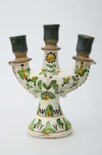 Obiect ceramic de uz casnic, format dintr-un picior tronconic, din care pornesc trei brațe, în care se introduc lumânările. Decorul pictural, realizat pe toată suprafața obiectului, este compus din motive geometrizante și vegetale. Primul registru, situat pe baza evazată, este format din elemente vegetale, dispuse circular și încadrat între două linii concentrice subțiri. Cel de-al doilea registru prezintă decor similar celui din registru inferior. Registrul superior este compus din trei flori, dintre care două (laterale) cu pistilul pronunțat, cea de-a treia, poziționată central, floarea soarelui. Gâtul și buza vasului au fost decorate cu verde și maro deschis. Iscălitură: indescifrabil. Cromatică: fond: alb; motive: crem; verde, maro închis, maro deschis, galben, negru.